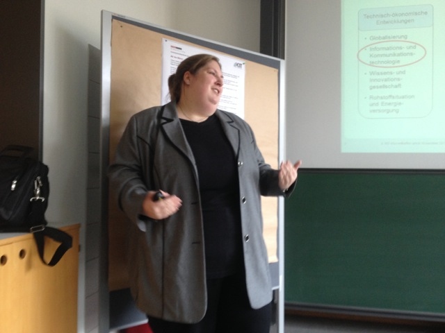 Frau im grauen Blazer referiert vor einer Präsentation zu technisch-ökonomischen Entwicklungen.